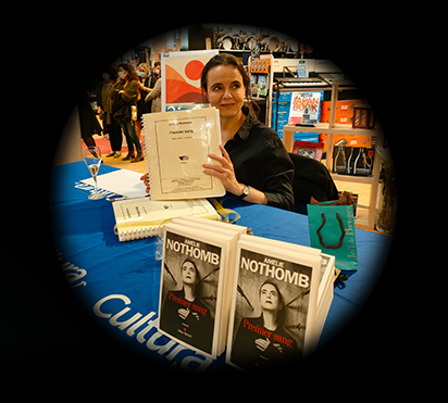Vue dans un cercle sur fond noir : Amélie Nothomb assise derrière une table, tenant son livre en braille.
