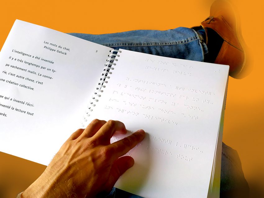 Un livre en braille et gros K, ouvert et posé sur des jambes croisées. Une main parcourt la page en braille.