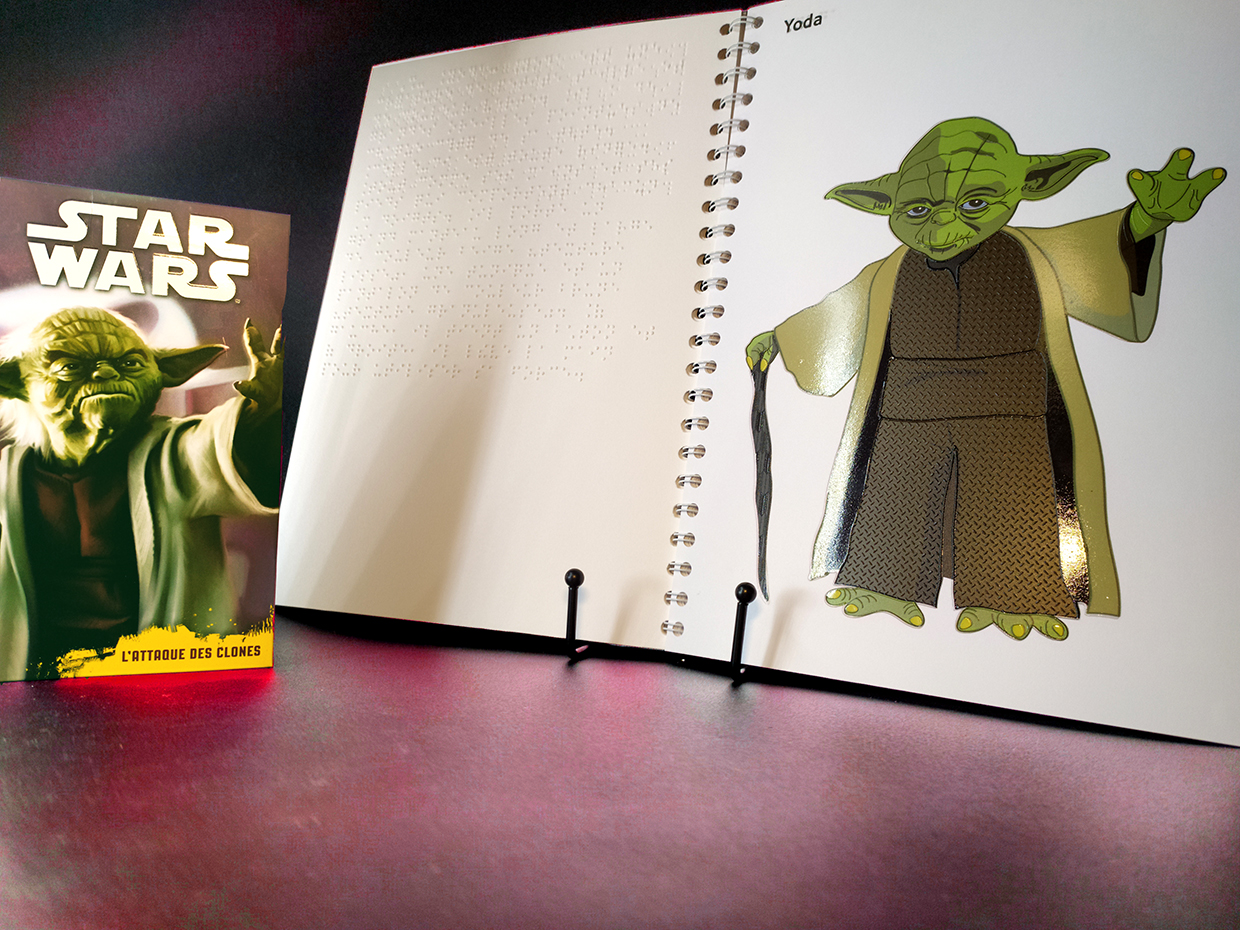 Une photo de maître Yoda devant le livre en braille de STAR WARS ouvert à la page qui comporte son illustration en relief.