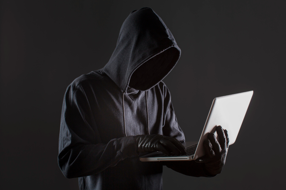 Une personne gantée et habillée de noir, visage caché sous une capuche, tient un ordinateur portable ouvert sur son avant-bras et pianote.